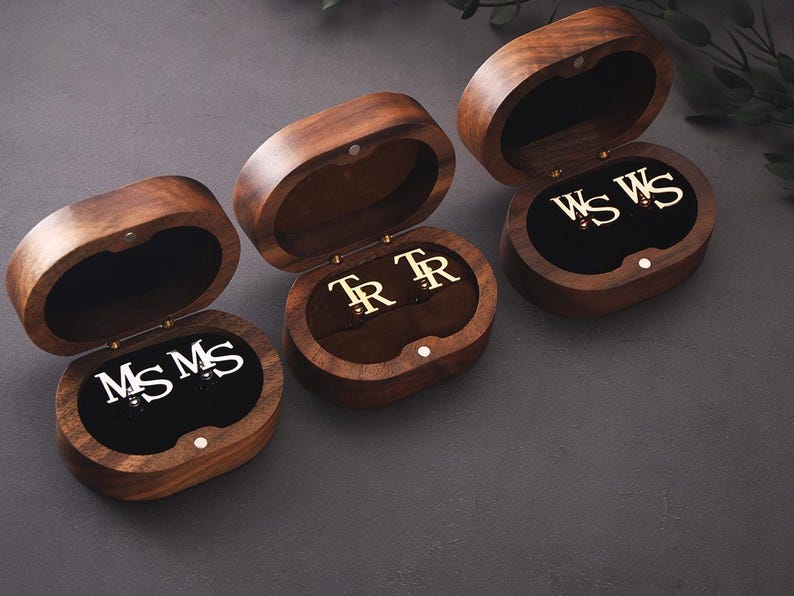 May include: Three open wooden jewellery boxes, each displaying a pair of initialled cufflinks. The boxes are oval, with a dark brown finish and a black velvet lining. The cufflinks feature silver or gold initials. The boxes are open.