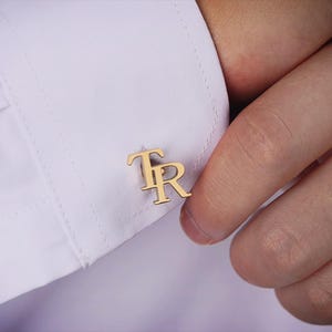 May include: A close-up of gold-coloured cufflinks bearing the initials "TR" on a white shirt cuff. The cufflinks are the focal point, presenting a classic, personalised accessory. The image emphasises the cufflinks' elegance.