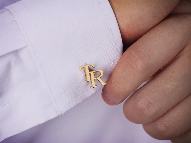 May include: A close-up of gold-coloured cufflinks bearing the initials "TR" on a white shirt cuff. The cufflinks are the focal point, presenting a classic, personalised accessory. The image emphasises the cufflinks' elegance.