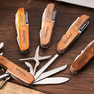 Könnte beinhalten: Holz-Multitools mit verschiedenen Werkzeugen, darunter Messer, Scheren und Flaschenöffner. Jedes Tool ist mit einem Namen und dem Text "GROOMSMAN 02.26.2025" graviert. Auf dunkler Holzoberfläche.