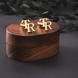 May include: Gold cufflinks, each with the letters "TR", rest on a dark wooden box. The box is a rich brown colour and sits against a dark grey backdrop. The cufflinks are a bright gold.