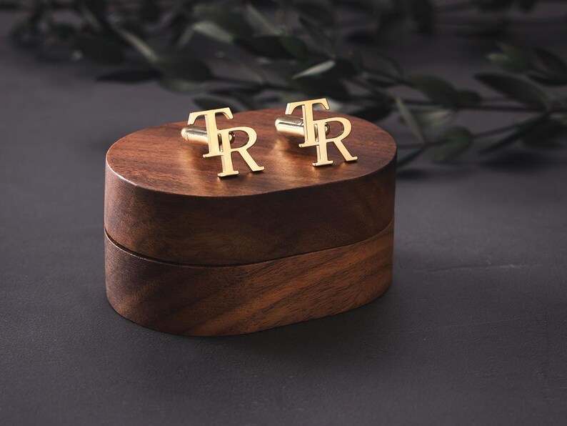 May include: Gold cufflinks, each with the letters "TR", rest on a dark wooden box. The box is a rich brown colour and sits against a dark grey backdrop. The cufflinks are a bright gold.