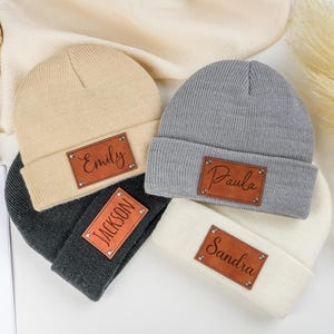 Könnte beinhalten: Vier Strickmützen in Beige, Grau, Dunkelgrau und Weiß, jede mit einem rechteckigen braunen Lederaufnäher. Die Aufnäher sind mit den Namen "Emily", "Paula", "Jackson" und "Sandra" personalisiert.