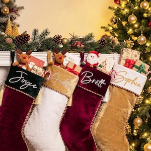 Könnte beinhalten: Vier Weihnachtsstrümpfe aus Samt und Plüsch mit den auf die Bündchen gestickten Namen Joyce, Aiden, Brett und Sophia. Die Strümpfe sind mit Plüsch-Rentier- und Weihnachtsmannfiguren, Zuckerstangen und kleinen Geschenkboxen verziert.