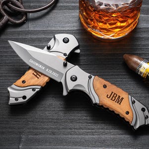 May include: A silver and wood folding knife with the text "GROOMSMAN 8.29.2022" and initials "H" engraved on the wood. The knife also has the initials "JBM" engraved on the handle. A cigar and a glass of amber liquid are in the background.