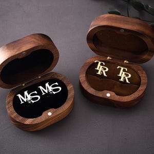 May include: Two open wooden jewellery boxes with oval shapes. One box contains silver initials "MS MS" on a black velvet interior. The other box has gold initials "TR TR" on a brown velvet interior. The boxes are on a dark grey surface.