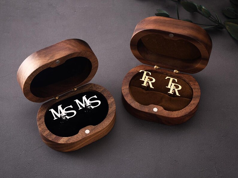 May include: Two open wooden jewellery boxes with oval shapes. One box contains silver initials "MS MS" on a black velvet interior. The other box has gold initials "TR TR" on a brown velvet interior. The boxes are on a dark grey surface.