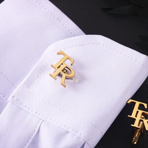 May include: Gold cufflinks featuring the letters "TR" on a white shirt cuff. The cufflinks are the focal point, showcasing their design. The shirt cuff is neatly folded, with a dark background.