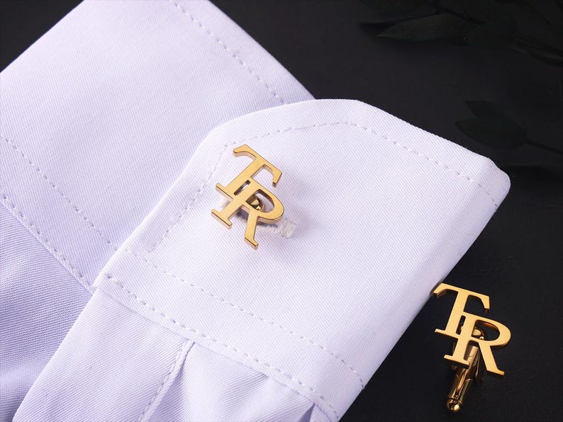 May include: Gold cufflinks featuring the letters "TR" on a white shirt cuff. The cufflinks are the focal point, showcasing their design. The shirt cuff is neatly folded, with a dark background.