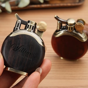 May include: Two vintage-style lighters with a round, dark wood body and brass accents. One lighter is held in a hand, displaying the engraved name "William" and the word "Groomsman". The other lighter is in the background.