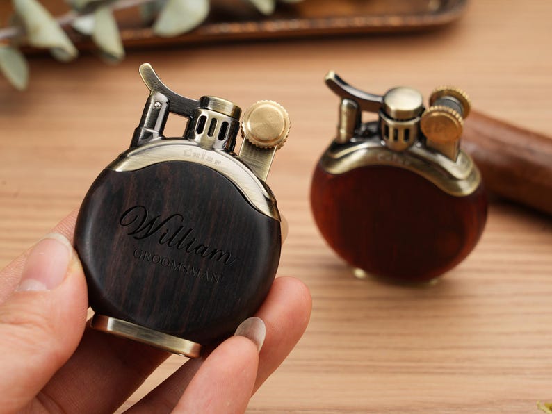 May include: Two vintage-style lighters with a round, dark wood body and brass accents. One lighter is held in a hand, displaying the engraved name "William" and the word "Groomsman". The other lighter is in the background.