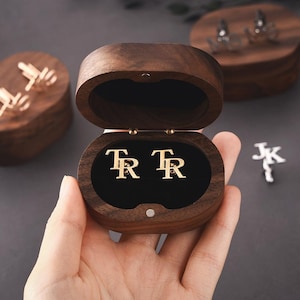May include: Gold cufflinks with the initials "TR" inside an open wooden box. Other cufflinks and wooden boxes are visible in the background. The background is a dark grey colour.