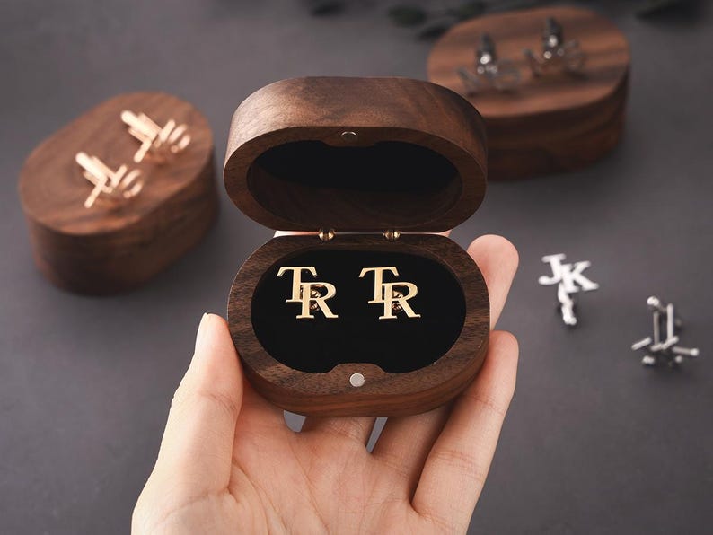 May include: Gold cufflinks with the initials "TR" inside an open wooden box. Other cufflinks and wooden boxes are visible in the background. The background is a dark grey colour.