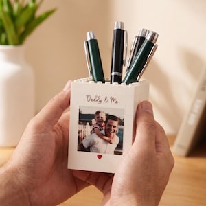 May include: A white pen holder with a photo of a father and child, the text "Daddy & Me", and a red heart. The holder contains several pens with black and green barrels. A decorative vase and books are in the background.