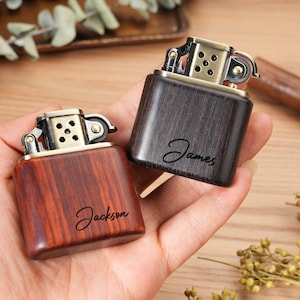 May include: Two wooden lighters with brass accents are held in a hand. One lighter is a reddish-brown color and the other is dark brown. The name "Jackson" is written on the reddish-brown lighter, and the name "James" is written on the dark brown lighter.