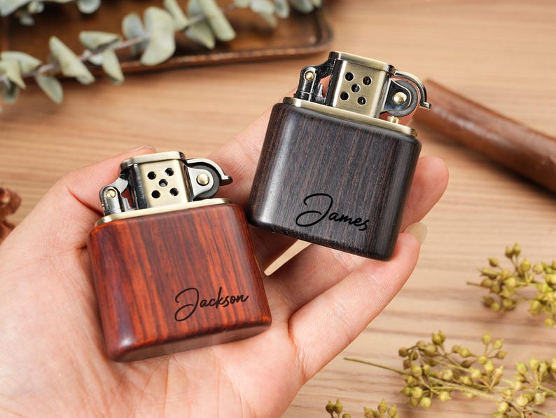 May include: Two wooden lighters with brass accents are held in a hand. One lighter is a reddish-brown color and the other is dark brown. The name "Jackson" is written on the reddish-brown lighter, and the name "James" is written on the dark brown lighter.