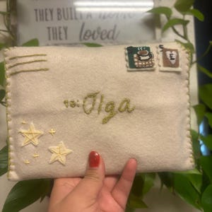 May include: Cream felt envelope with embroidered details. Features two star appliqués, two postage stamp appliqués, and the embroidered text "to: Olga". The envelope is stitched with green thread.