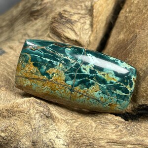 May include: A polished, rectangular gemstone with a mix of teal, green, and brown hues. The stone has a glossy finish and features intricate veining patterns throughout its surface. The gemstone is a natural stone, possibly a type of jasper.