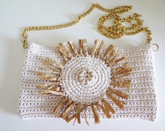 Puede incluir: Un bolso de mano blanco de crochet con una correa de cadena dorada. El bolso presenta un diseño de sol tejido marrón en el centro.