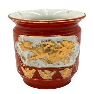May include: A red ceramic pot with a gold dragon design on a light blue background. The pot has a white interior and gold trim. The bottom of the pot has gold accents. This decorative pot is a unique piece.