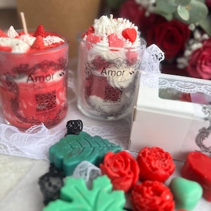 May include: Two glass candles with red and white layers, topped with whipped cream and fruit. The candles have the text "Amor Achille Lauro" on the glass. Also, there are red rose-shaped wax melts and green leaf-shaped wax melts.