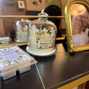 Puede incluir: Una vela de campana de cristal con base blanca, llena de una vela perfumada y flores decorativas. La vela tiene una etiqueta con texto italiano. Un paquete de ceras perfumadas está en primer plano.