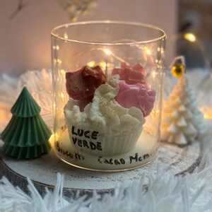 Pode incluir: Um castiçal de vidro transparente com uma vela em forma de cupcake no interior, coberta com cera em forma de rosa vermelha e rosa. A vela é rodeada por luzes de fadas e assenta numa base de madeira. O texto "LUCE VERDE" está escrito no vidro.