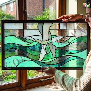 Puede incluir: Un panel rectangular de vidriera que presenta una representación estilizada de piernas sumergidas en capas onduladas de vidrio en tonos verdes y azulados. La pieza está enmarcada en madera oscura e incluye el texto COLORLYBYVN en el centro.