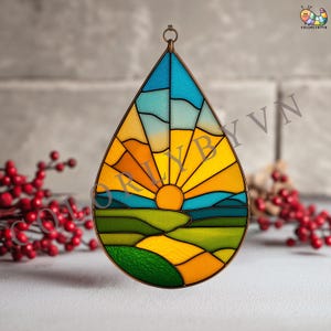 May include: A stained glass suncatcher in a teardrop shape, depicting a sunrise over green hills. The design uses blue, yellow, and orange hues, with a copper-coloured metal frame and a hanging loop. The text "COLORLYBYVN" is visible.