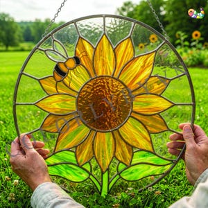 May include: A round stained glass window featuring a sunflower with yellow and orange petals and a brown center. A black and yellow bee is on the left side. Green leaves are at the bottom. The window is held by hands.