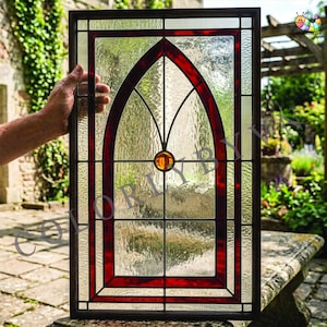 May include: A rectangular stained glass window panel featuring a dark red arched frame and a central amber glass cabochon. The panel is constructed with textured clear glass panes and a dark metal border, showcasing a classic gothic architectural design.