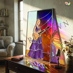 May include: Stained glass panel depicting a woman playing a guitar. The artwork features a color palette of purple, orange, and blue. The woman is wearing a purple dress. The panel is displayed on a wooden table with a vintage microphone and a vase of flowers.