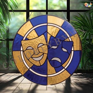 May include: Round stained glass art piece featuring the comedy and tragedy theater masks. The masks are gold and blue, set against a gold and blue background. The piece is designed to hang, with a chain at the top.