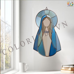 Może przedstawiać: Witraż przedstawiający postać religijną z niebieskim nimbem i szatą. Dzieło sztuki zawiera odcienie niebieskiego, zielonego i beżowego, z ramą w kolorze miedzi. Obraz wisi na białej ścianie, w tle okno i kubek.