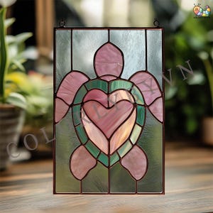 以下が含まれることがあります： ピンクとグリーンのウミガメのデザインが特徴のステンドグラスパネル。ハート型の甲羅が特徴です。長方形のパネルは、濃い茶色のフレームと吊り下げ用のループが付いています。カメの甲羅はピンクとグリーンのグラデーションになっています。