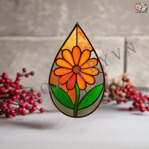 May include: A stained glass suncatcher in a teardrop shape, featuring an orange daisy with green leaves and a brown border. The background is a mix of orange and gray glass, with a small loop for hanging.