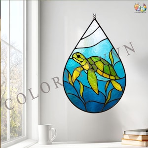 May include: A stained glass window in a teardrop shape, featuring a sea turtle swimming in blue water with yellow and green accents. The window is framed in black and hangs against a white wall.