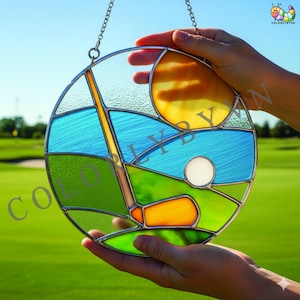 May include: A round stained glass golf scene with a golf club, ball, and sun. The artwork features blue, green, yellow, and orange colors. The glass is held by a silver chain.