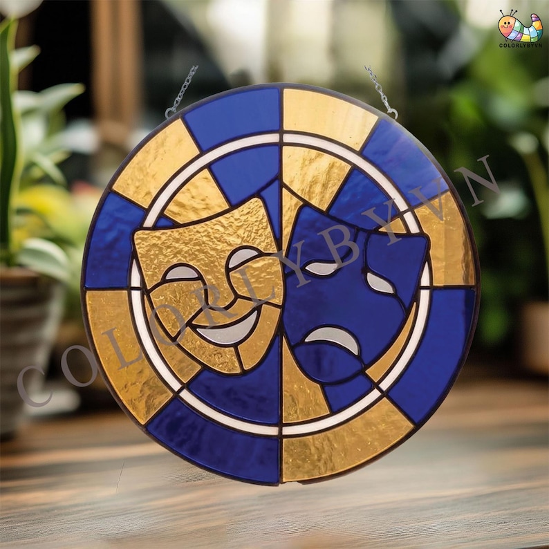 May include: Round stained glass art piece featuring the comedy and tragedy masks. The design incorporates gold, blue, and white colors. The masks are set against a gold and blue checkered background, with a white border. The piece is suspended by a chain.