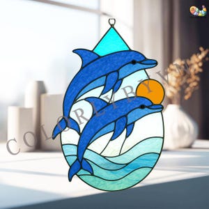 Op de afbeelding: Gebrandschilderd glazen zonnevanger met twee blauwe dolfijnen die boven oceaangolven springen, met een oranje zon op de achtergrond. De zonnevanger heeft een druppelvorm met een turquoise driehoek aan de bovenkant. Het woord "COLOR" is zichtbaar.