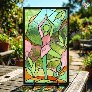 May include: Stained glass panel depicting a figure in a yoga pose, framed in black. The figure is pink and green, surrounded by green and orange leaves against a light blue and green background. The artwork is displayed on a wooden table.