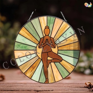 May include: A round stained glass piece featuring a yoga pose. The design includes a figure in a tree pose, surrounded by a sunburst pattern in shades of green, yellow, and brown. The text "COLOR BY YIN" is visible.