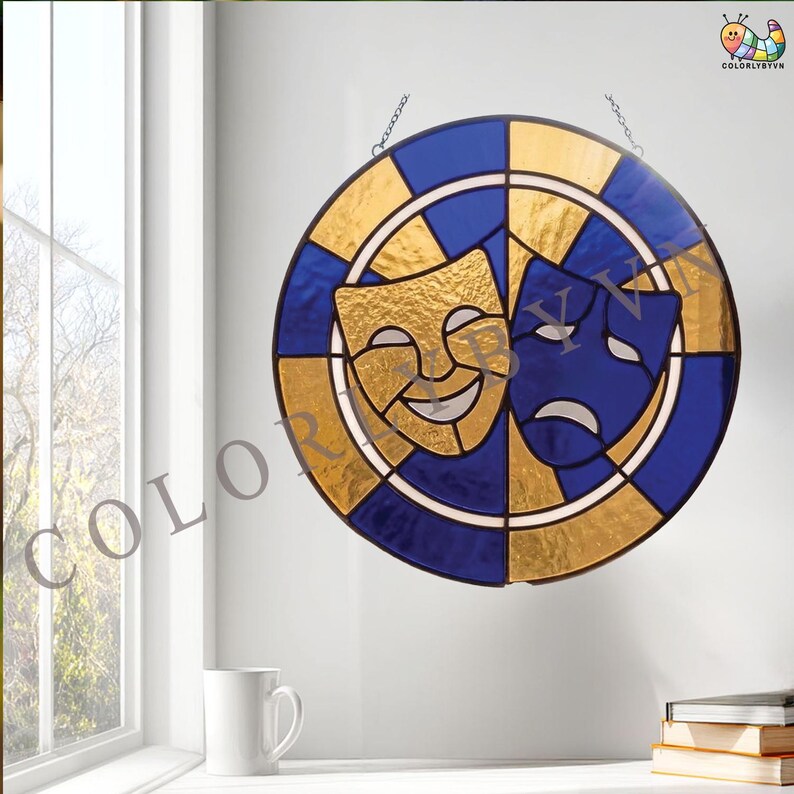 May include: Stained glass art piece featuring the comedy and tragedy theater masks. The round design has a gold and blue color scheme. The masks are gold and blue, with a white background. The piece is hanging from a chain.