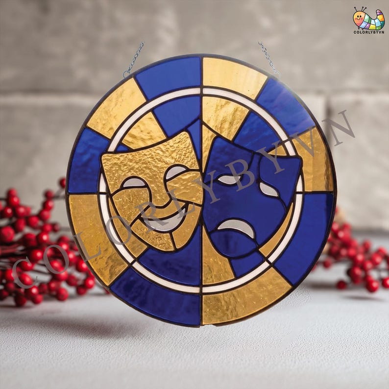 May include: A round stained glass piece featuring the comedy and tragedy theater masks. The masks are gold and blue, set against a gold and blue background. The piece is designed with a circular pattern and has a chain for hanging.