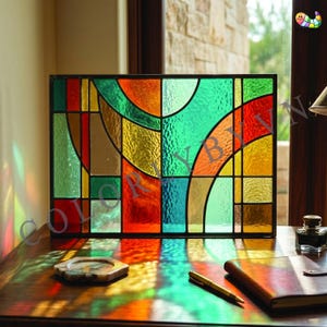 May include: A rectangular stained glass panel with a black frame. The panel features an abstract design with sections of green, orange, red, yellow, and brown glass. The text "COLOR BY" is visible on the glass. The panel is on a wooden surface.