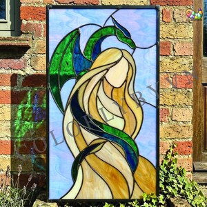 Può includere: Pannello in vetro colorato raffigurante una donna con lunghi capelli biondi e un drago verde e blu. L'opera d'arte ha una cornice nera e uno sfondo azzurro. Le ali del drago sono di un verde vibrante.