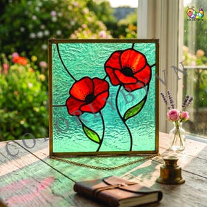 May include: Stained glass panel depicting two red poppy flowers with black centres and green stems against a textured turquoise background. The panel is framed in gold and hangs by a delicate chain. A notebook and small brass bell sit on a wooden surface.