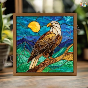 May include: Stained glass artwork featuring an eagle perched on a branch, set against a backdrop of a yellow sun, blue sky, and mountains. The eagle has brown and white feathers. The artwork is framed in a wooden frame.