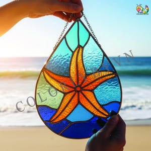 May include: A stained glass window in a teardrop shape, featuring a starfish design. The starfish is orange and red, set against a backdrop of blue and green glass. A metal chain is attached for hanging. The background shows the ocean and sky.