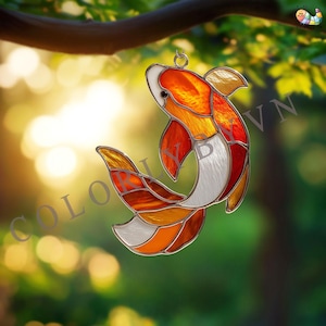 Peut inclure: Suncatcher en vitrail représentant une carpe koï aux couleurs orange, rouge, blanc et jaune. Le poisson est suspendu à une boucle métallique. Le texte "COLOR BY VN" est visible. Fond vert flou.